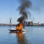 Lancha em chamas no Lago Paranoá, Brasília, com fumaça e reflexos na água, representando incêndio que causou queimaduras graves.