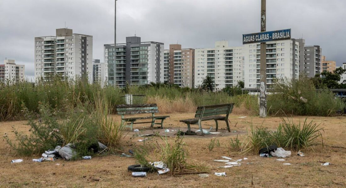 Área verde negligenciada em Águas Claras, com vegetação seca, lixo e bancos quebrados, ao fundo prédios modernos de Brasília.