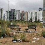 Área verde negligenciada em Águas Claras, com vegetação seca, lixo e bancos quebrados, ao fundo prédios modernos de Brasília.