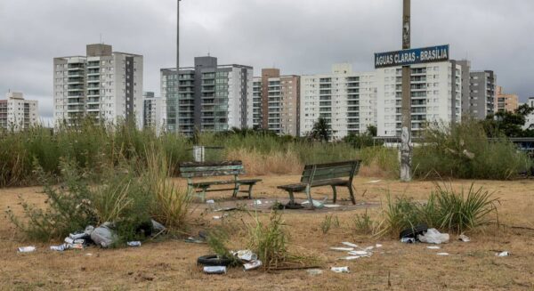 Área verde negligenciada em Águas Claras, com vegetação seca, lixo e bancos quebrados, ao fundo prédios modernos de Brasília.