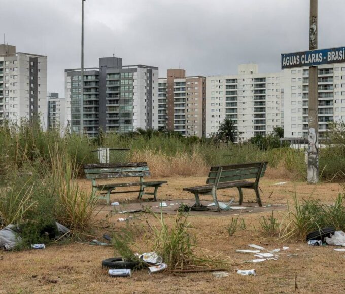 Área verde negligenciada em Águas Claras, com vegetação seca, lixo e bancos quebrados, ao fundo prédios modernos de Brasília.